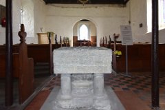 Vange - All Saints - Font  View of the 12th century font taken from under the gallery at the west end of the church. Its square bowel of Purbeck marble, which has a crudely incised zigzag pattern on its eastern face, rests on a circular central pillar and four more recently renewed smaller columns. The four corners of the bowl have worn trefoil decorations and in the north east and south west corners are traces of the locking device to prevent baptismal water being stolen. : Church, Essex, Vange, All Saints, Norman, Font